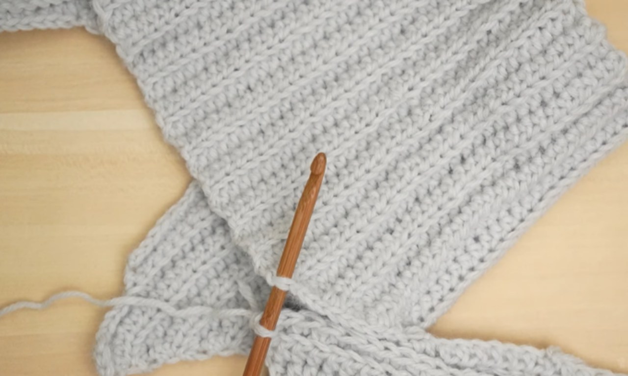 A close-up of a light gray, ribbed crochet project laid flat on a wooden table, with a wooden crochet hook inserted into a stitch to demonstrate a part of the pattern.