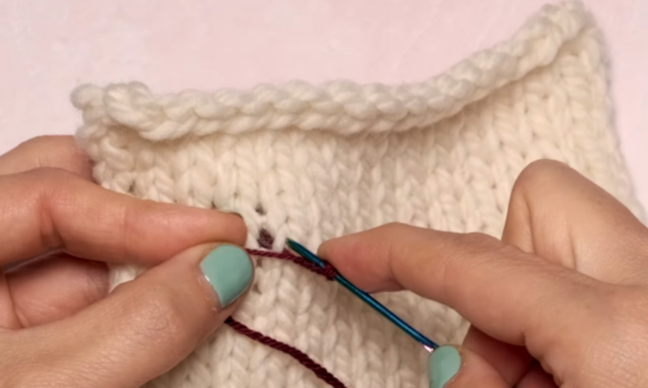 A person's hands demonstrating how to embroider on knit fabric. They are holding a piece of cream-colored knitwear and guiding a blue darning needle with dark red thread to make a stitch.