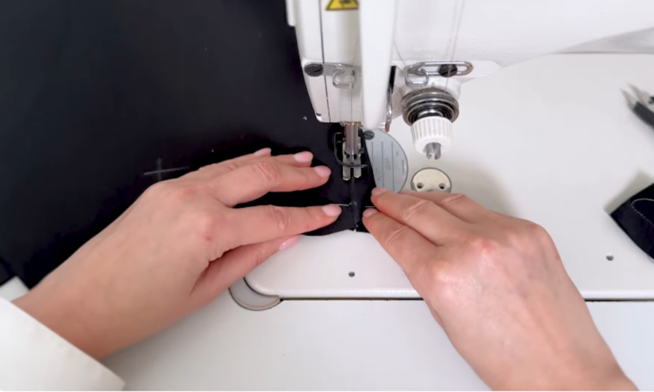 A close-up of hands sewing a dart on black fabric for the drop-waist dress bodice, guiding it carefully under the needle of a sewing machine.