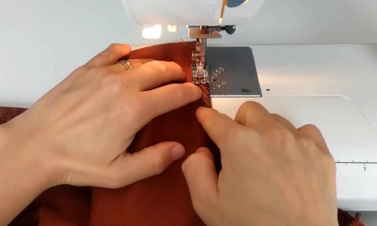 A close-up of a person's hands carefully guiding two layers of burnt orange fabric under the presser foot of a sewing machine to sew a seam.