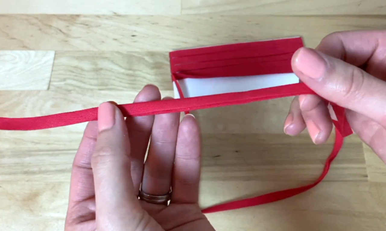 A close-up of a person's hands holding a length of red, single-fold bias tape, which is unwound from its white cardboard packaging over a wooden surface.
