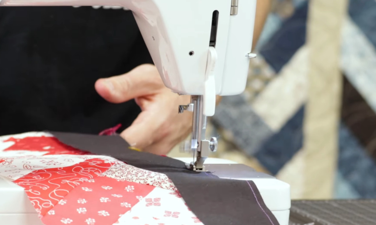 A close-up of a sewing machine stitching a black fabric border onto a red and white patterned French braid quilt piece, with a hand guiding the fabric.