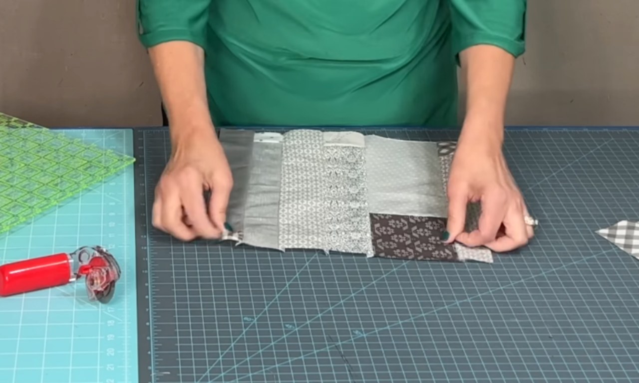 A person wearing a green shirt is working on a quilting project at a crafting table. They are smoothing out a long strip of pieced-together fabric made from various grey, black, and white patterned swatches. The fabric strip is laid across a large, aqua-colored gridded cutting mat. To the left on the mat are a red rotary cutter and a large, transparent green quilting ruler.