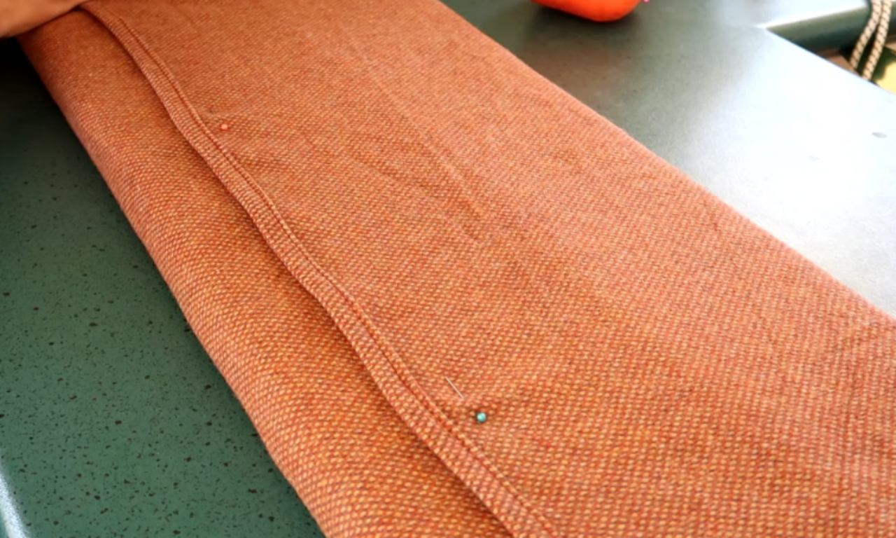 A close-up of a textured orange fabric cushion cover being pinned for sewing. The fabric is folded on a green surface, and a single pin with a turquoise head holds the hem in place.