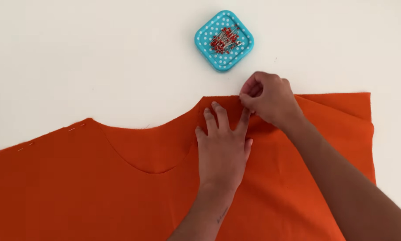 A sewer pinning the shoulder seams of the orange boxy top together on a white table, with a blue polka-dot pin cushion nearby.