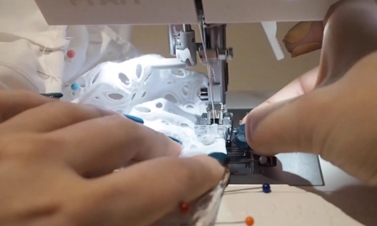 A sewer's hands carefully guiding white eyelet lace fabric through a sewing machine to stitch a French seam.