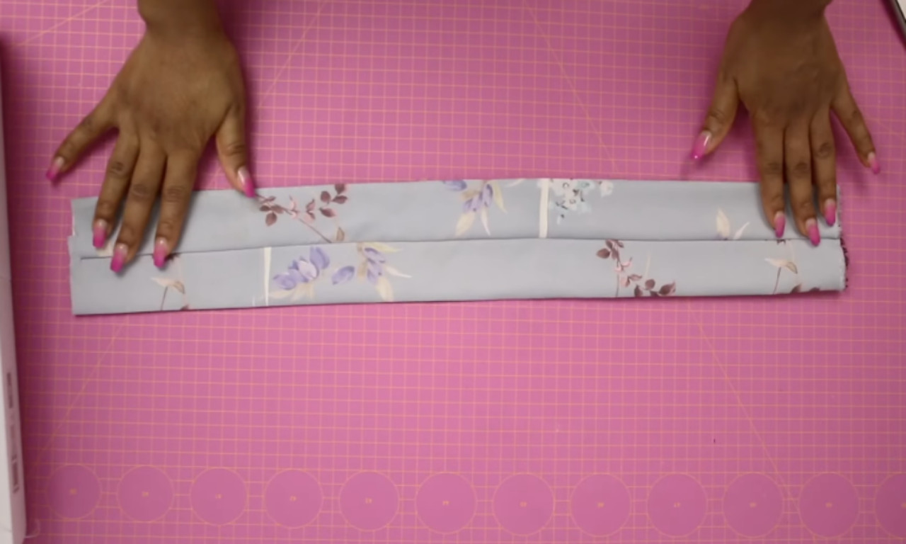 A sewer's hands holding a long piece of light blue floral fabric that has been folded lengthwise on a pink cutting mat, demonstrating a step in how to sew a knot headband.