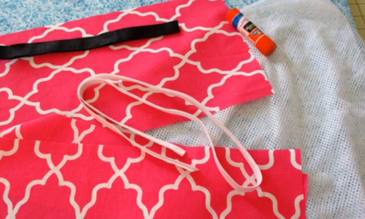 Sewing supplies for a DIY mesh laundry bag laid out on a table, including pink patterned fabric, white mesh, bias tape, and a glue stick.