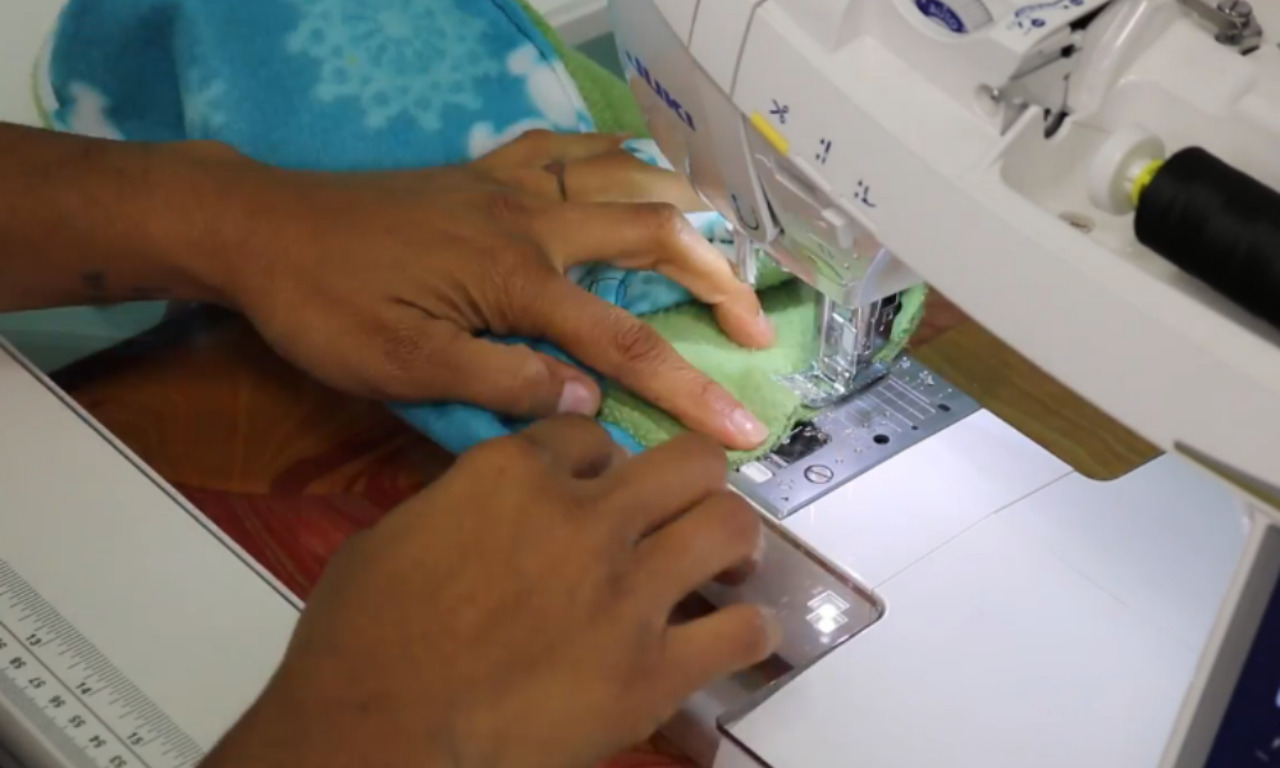 A close-up of hands guiding a blue and green fleece neck warmer under the needle of a white sewing machine to sew the final circular seam.
