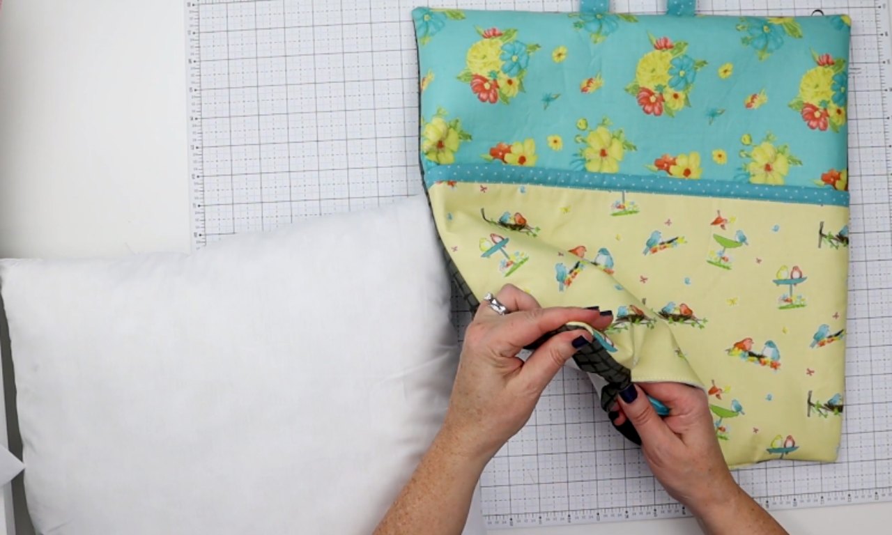 A person uses a blue point turner tool to smooth the seams inside a newly sewn reading pillow cover. The colorful cover sits on a gridded cutting mat next to a white pillow form, ready for stuffing.