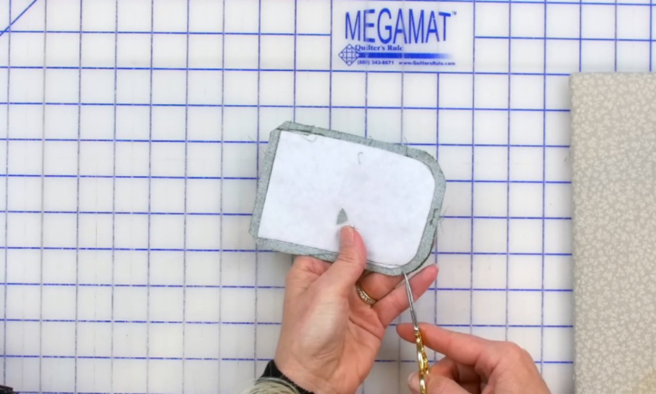 A person using small scissors to clip the curved seam allowance on an inside-out fabric gift tag, which is placed on a white and blue gridded sewing mat.
