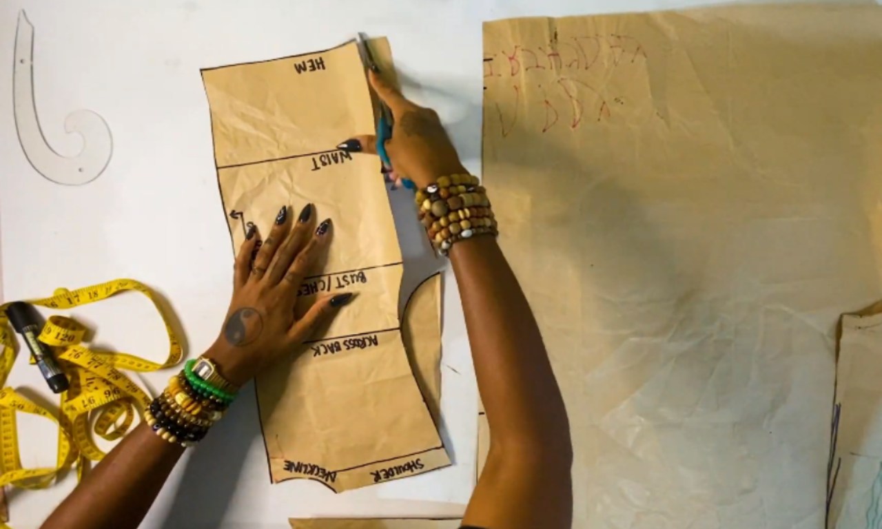 A person drafting a sewing pattern by cutting out a paper bodice piece labeled with "hem," "waist," and "bust." Sewing tools, including a measuring tape and French curve, are visible on the work table.