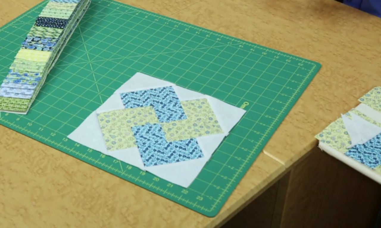 A completed Glad All Over quilt block laid out on a green cutting mat, showing the woven pattern of blue and yellow fabrics. A stack of jelly roll strips is in the background.