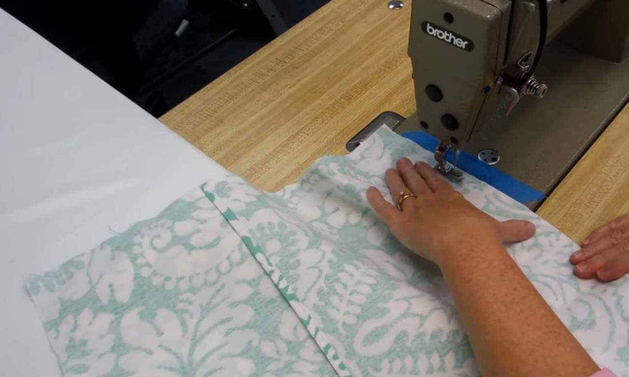 A person guides the patterned fabric under a sewing machine needle to stitch the seams of the envelope pillow cover.