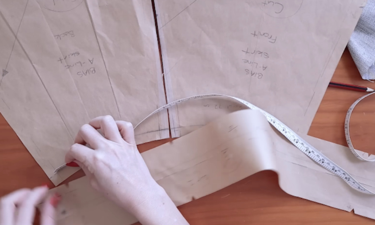 A sewer measures the curved waistline of a brown paper skirt pattern with a tape measure to get an accurate length before sewing the waistband.