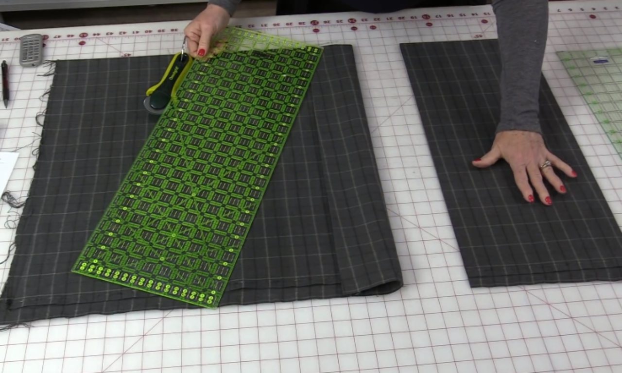 A quilter using a long green ruler and a rotary cutter to cut a strip of gray plaid flannel fabric on a gridded cutting mat.