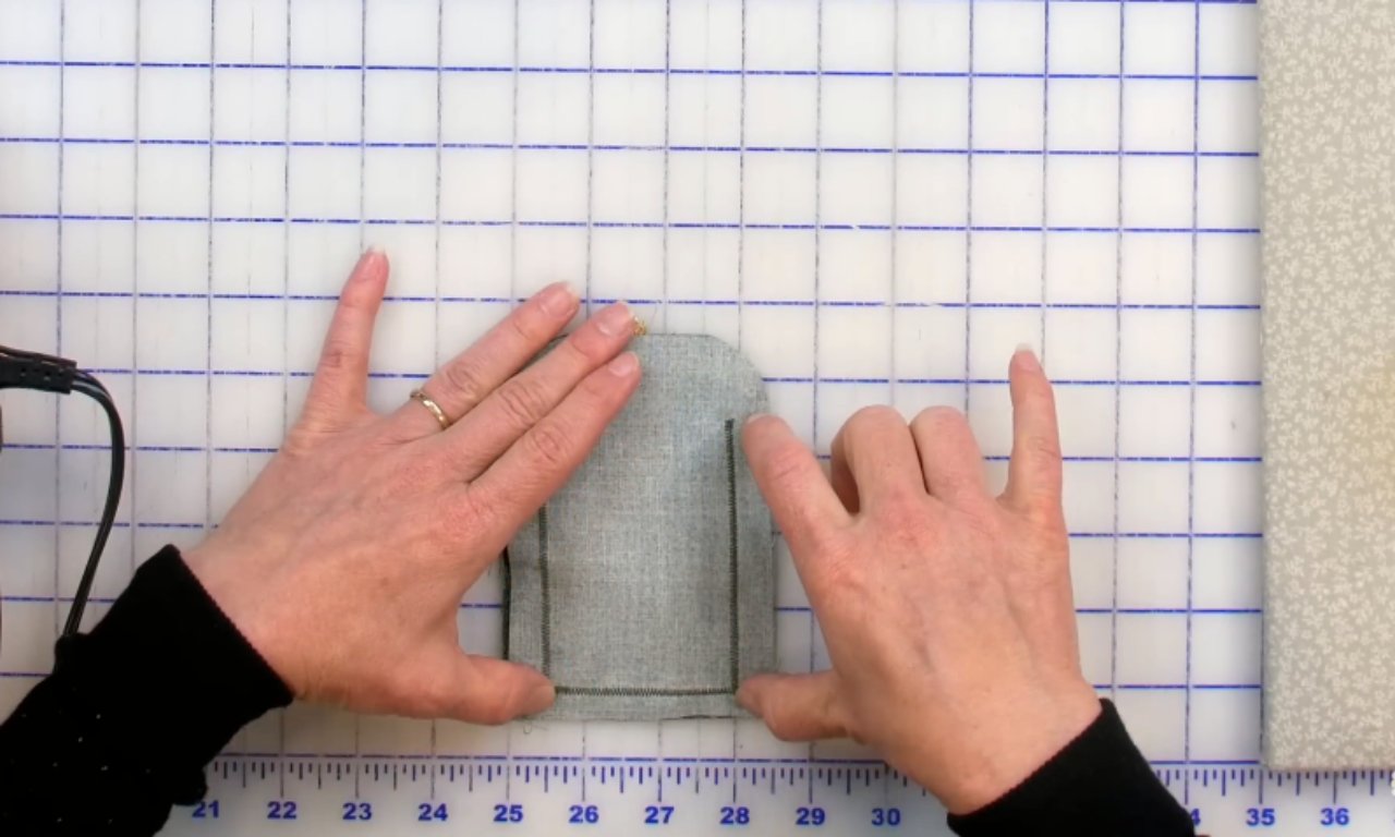 Hands holding the backing piece of a fabric gift tag on a gridded sewing mat, showing the U-shaped stitching that forms the pocket.