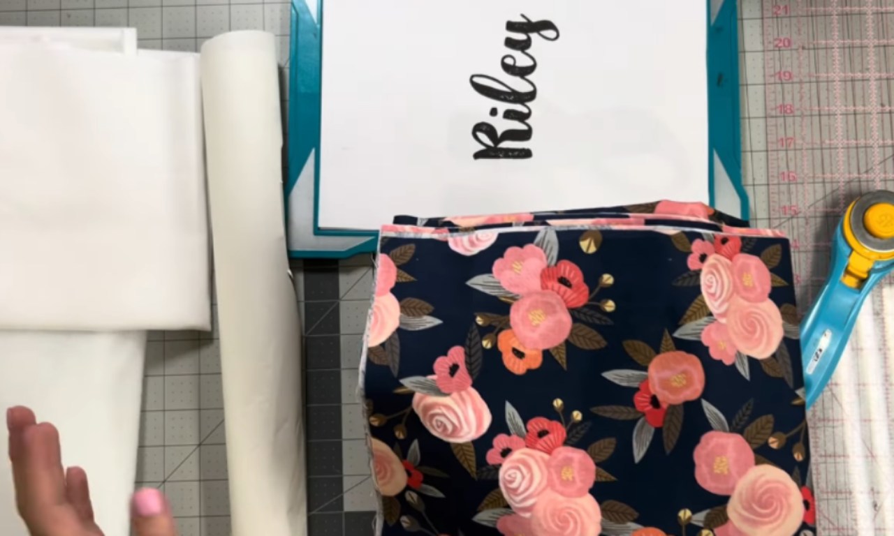 An overhead view of supplies for an appliqué quilt laid out on a cutting mat, including white fabric, floral fabric, batting, a rotary cutter, and a paper pattern with the name "Riley" written in script.