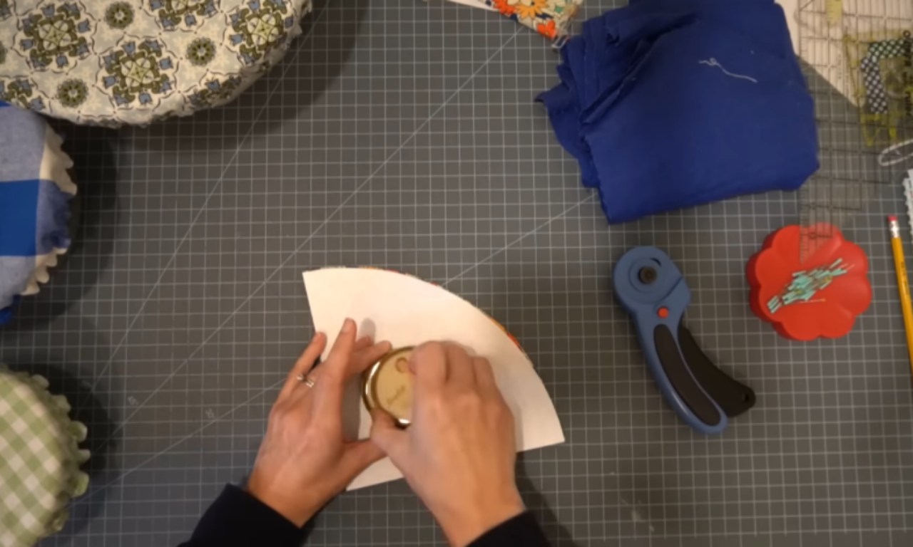 A top-down view of a person's hands using a jar lid to trace a rounded corner onto a quarter-circle paper pattern. The pattern is on a grey gridded cutting mat, surrounded by sewing supplies like a rotary cutter and finished cloth bowl covers.