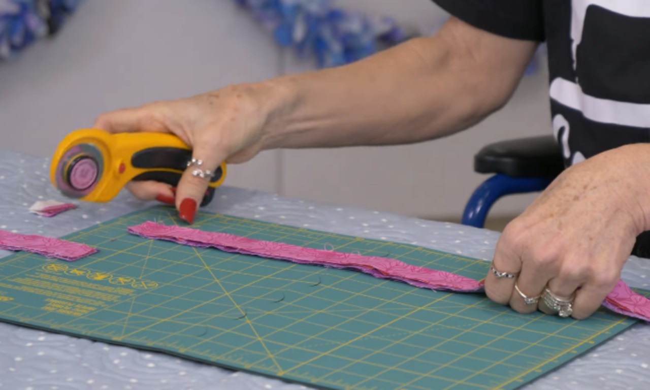 A person's hands using a yellow rotary cutter to cut a long strip of pink patterned fabric on a green cutting mat, preparing the strips for a DIY fabric garland.