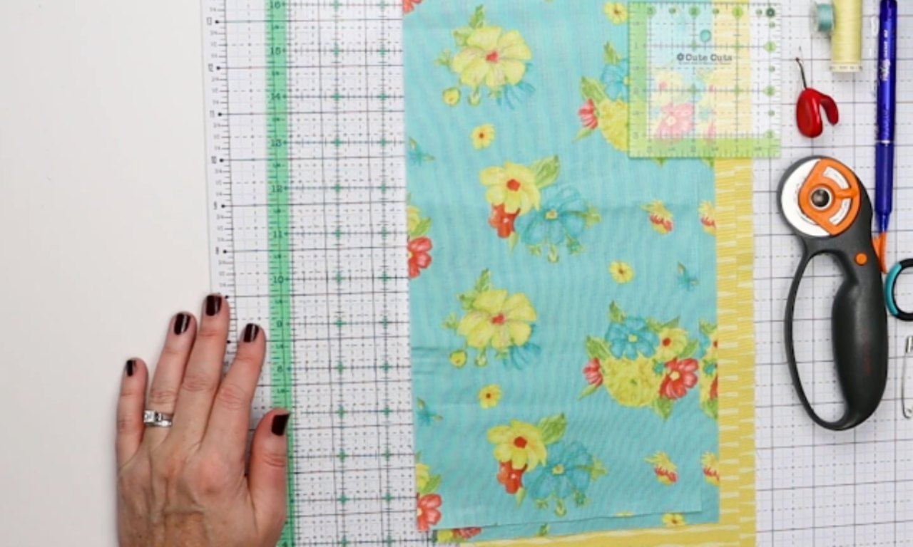 An overhead view of the sewing supplies needed for a DIY drawstring wine bag, including blue floral fabric, a ruler, rotary cutter, and thread laid out on a cutting mat.
