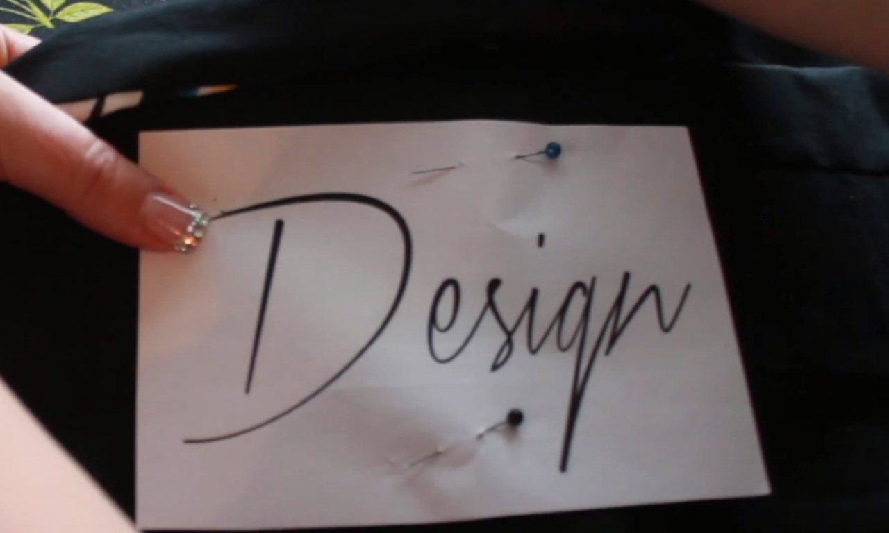 A close-up of a hand pinning a paper pattern to dark fabric. The white paper has the word "Design" printed in a black, cursive font and is held in place by two sewing pins, preparing for an embroidery project.