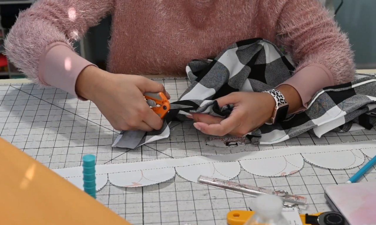 A sewer uses small scissors to carefully clip the corner of a sewn scalloped hem on black-and-white plaid fabric, with the paper pattern piece visible on the cutting mat.