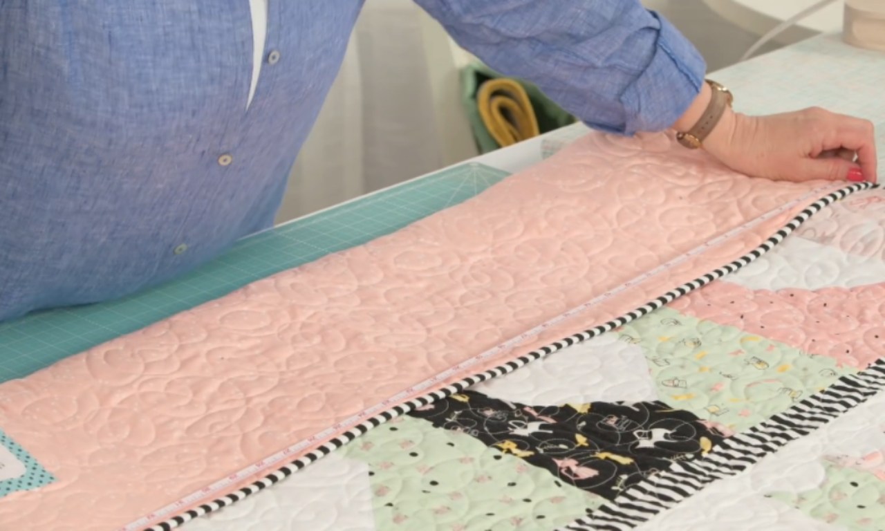 A woman in a blue shirt measures the edge of a colorful patchwork quilt with a tape measure on a cutting mat.