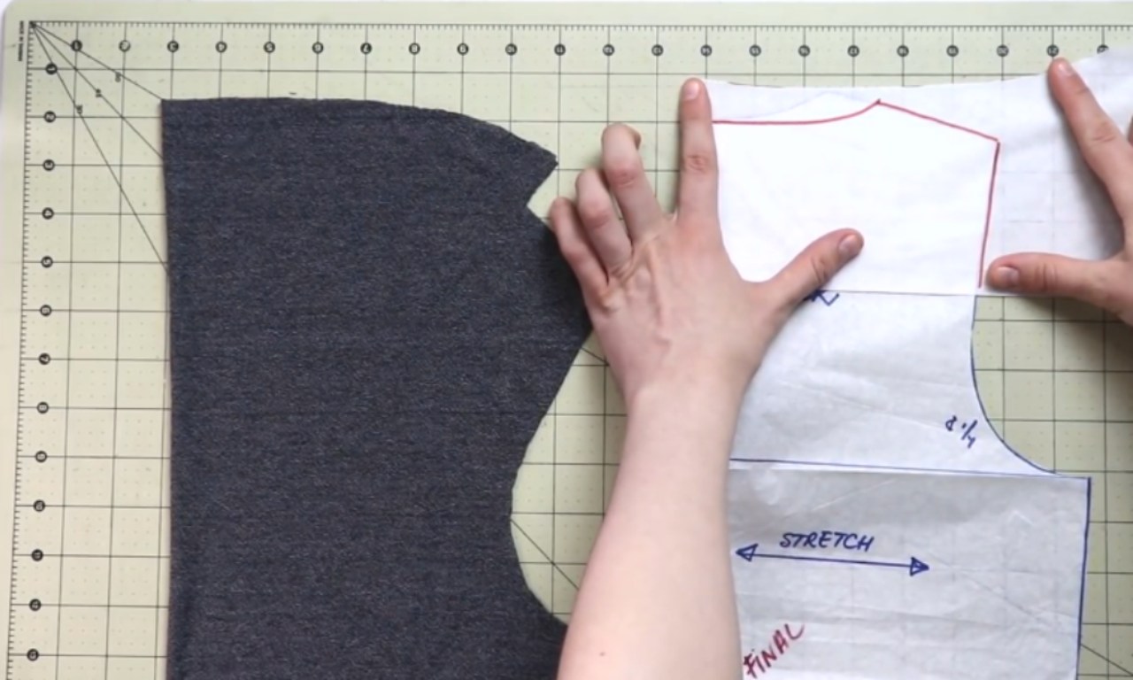A person's hands align a paper sewing pattern on a gridded cutting mat next to a piece of dark gray fabric.