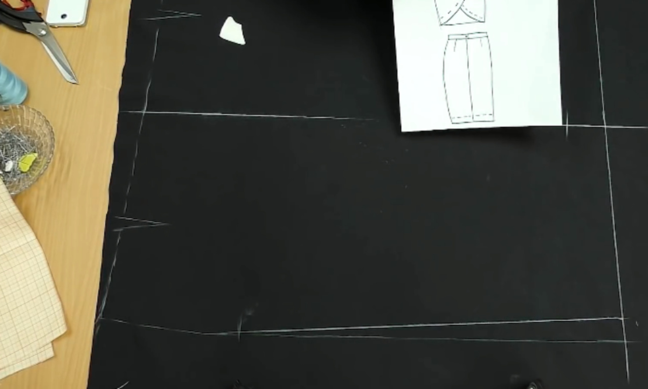 Drafting a tulip skirt pattern with white chalk directly onto black fabric laid out on a table. A paper sketch of the skirt design is visible for reference.