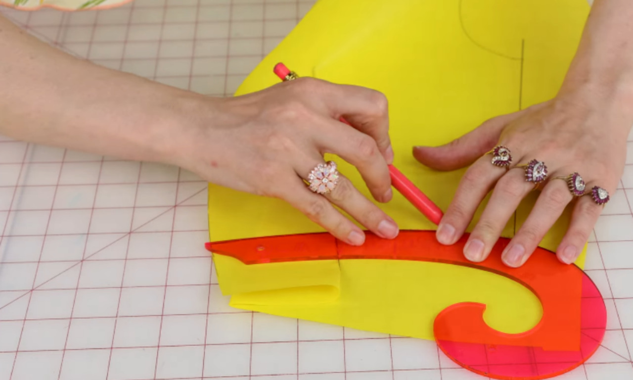 A person uses a pink pencil and an orange French curve ruler to draw a line on a piece of yellow paper on a gridded cutting mat.