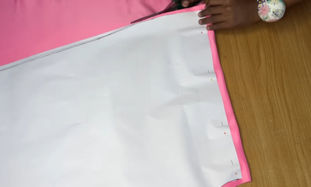 A sewer's hands cutting bright pink fabric on a wooden table, using scissors to follow the edge of a large white paper pattern pinned on top.