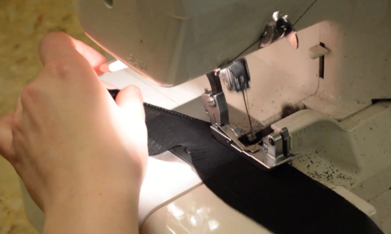 A hand guides black fabric through a serger sewing machine to create a rolled hem for the DIY corset belt's ruffle trim.