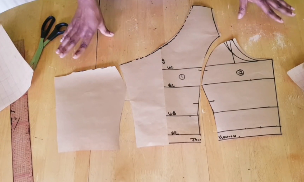 The drafted paper pattern pieces for a DIY one-shoulder top laid out on a wooden table, with a sewer's hands gesturing to the different components.
