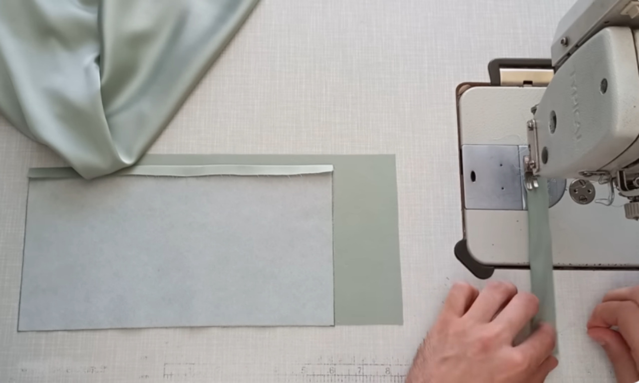 A sewer prepares a light green sleeve cuff by applying fusible interfacing, while sewing a narrow fabric strip for a button loop on a sewing machine.