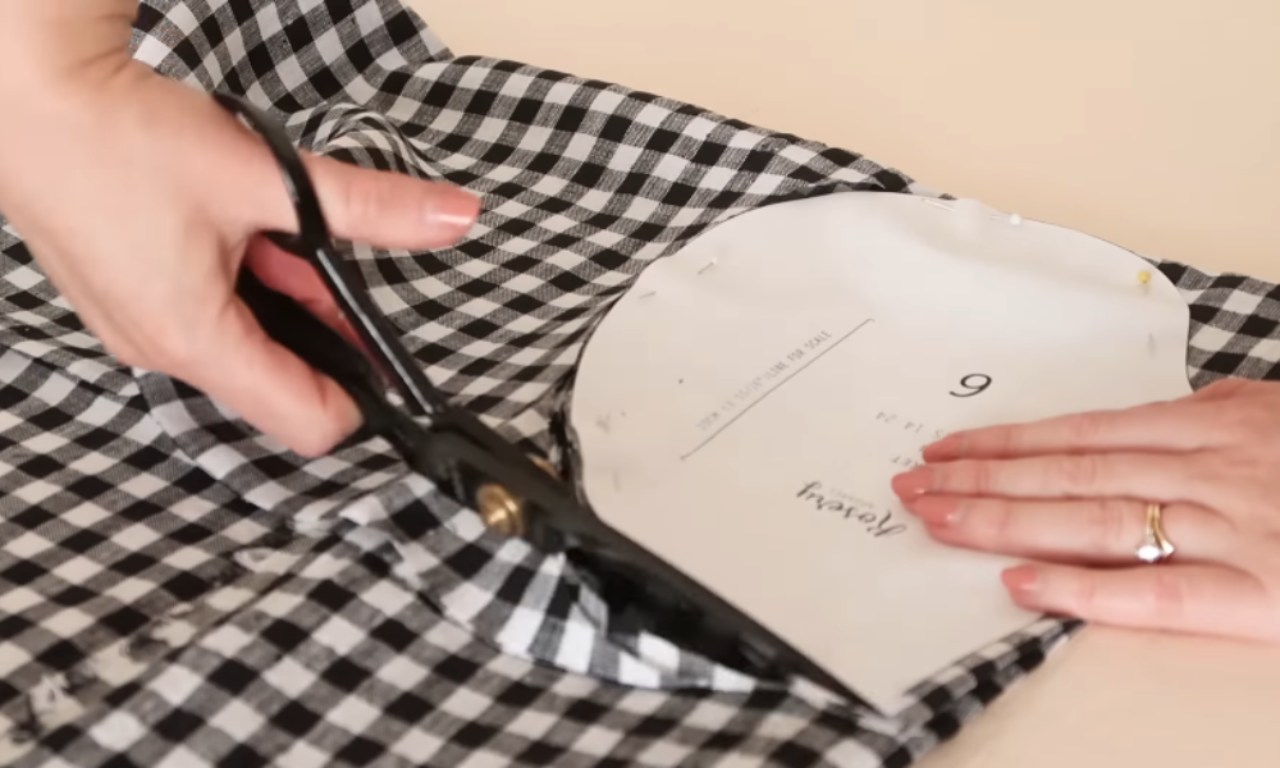 A person cutting black and white gingham fabric with scissors, using a paper sewing pattern as a guide.