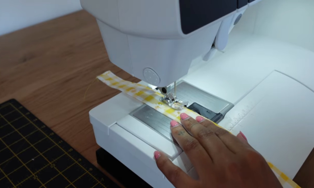 A hand guiding a yellow and white gingham fabric strap under the presser foot of a sewing machine to topstitch the edge.