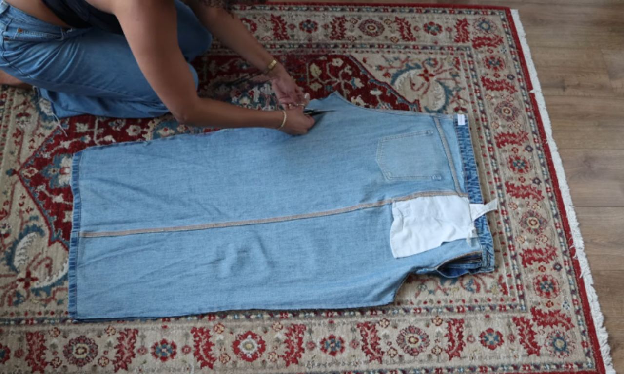 A person carefully cuts the curved crotch seam of a pair of jeans laid flat on a rug, a key step in the DIY process of turning jeans into a skirt.