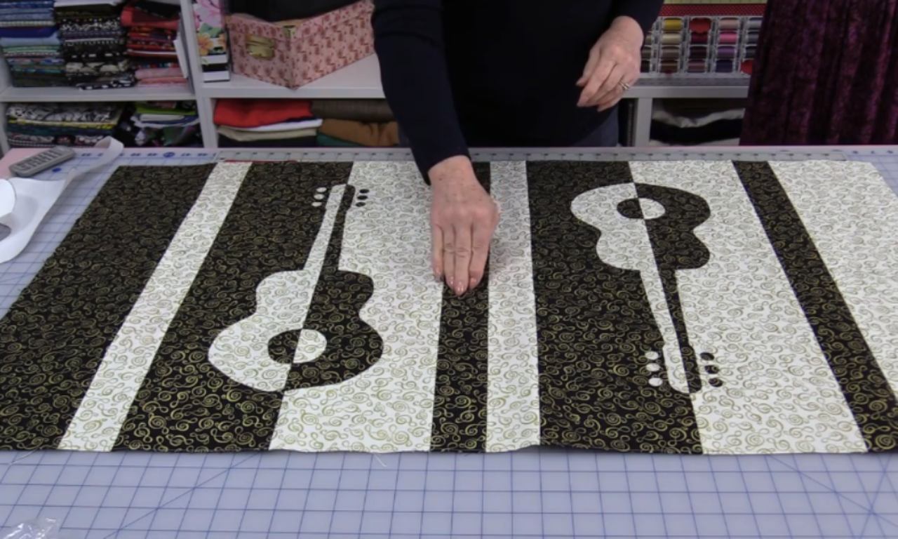 A person displaying a finished black and cream table runner with a reverse applique guitar design. The runner features two guitars that are mirror images of each other, one cream on a black background and the other black on a cream background, both with a gold music note pattern.