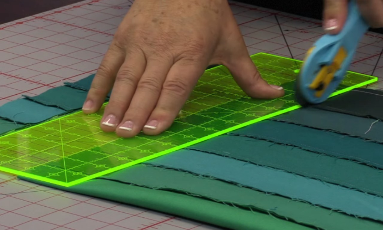 A quilter's hand holds a green acrylic ruler down on a sewn tube of teal and green fabric strips, carefully slicing it with a rotary cutter to create a new strip for an easy bargello quilt pattern.