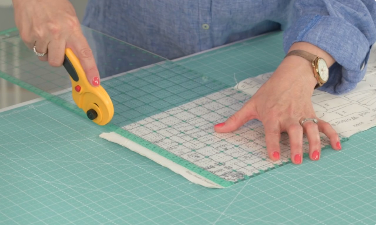 A person uses a yellow rotary cutter and a clear ruler to cut fabric on a green gridded cutting mat.