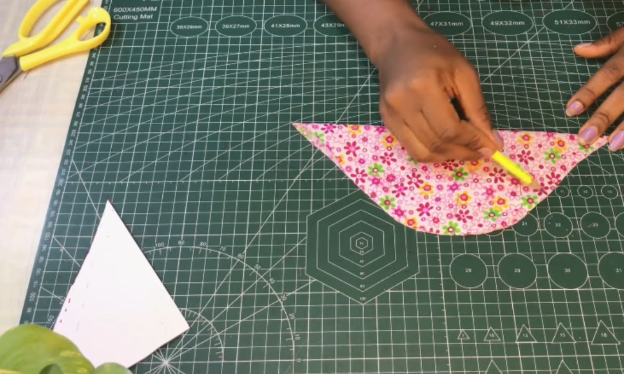 A person marks a pink floral fabric, cut in the shape of a cap sleeve, with yellow tailor's chalk on a green cutting mat.