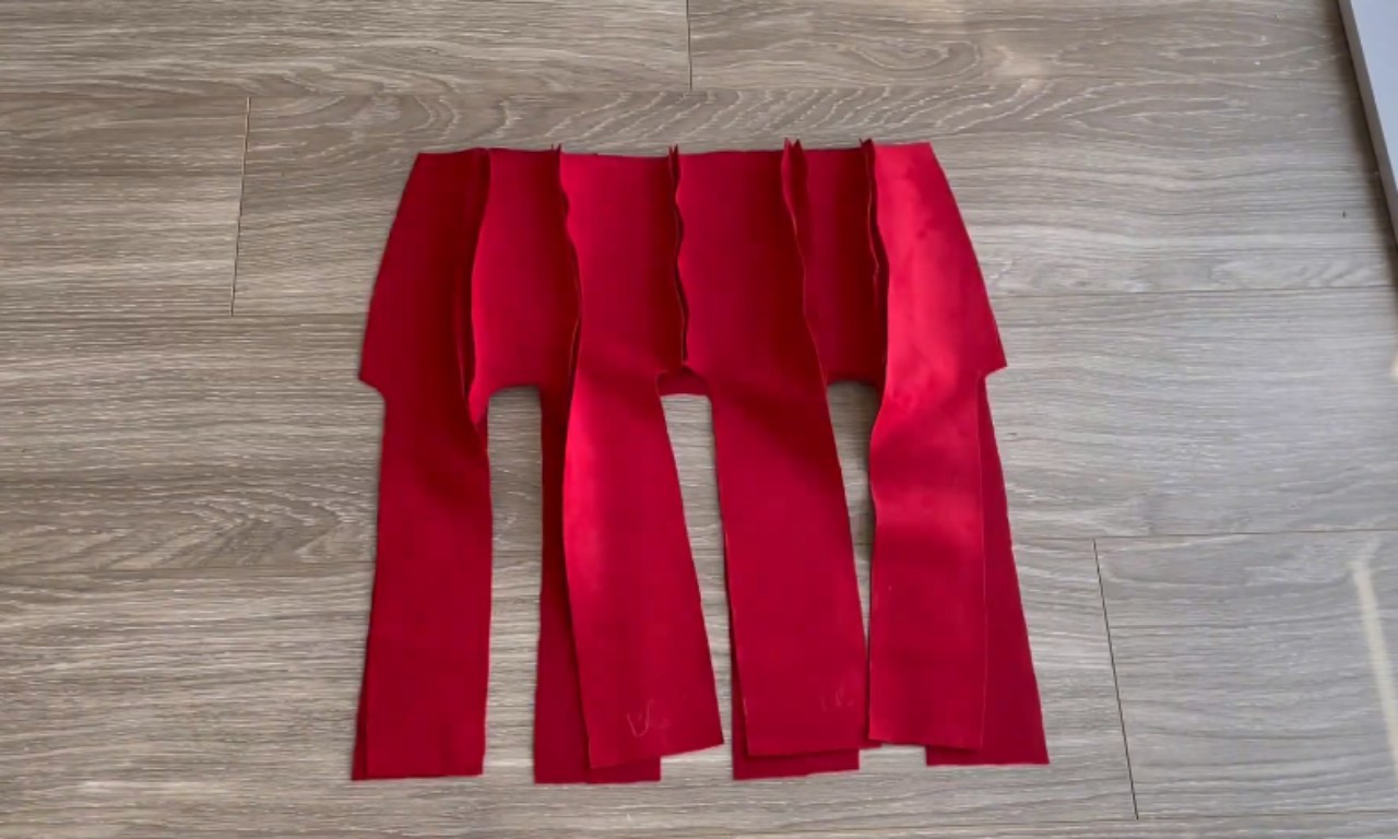 The red panels of a skirt are shown partially assembled on a wooden floor. The panels are sewn together at the top hip section, while the lower parts remain open, creating long slits where godets will be inserted.