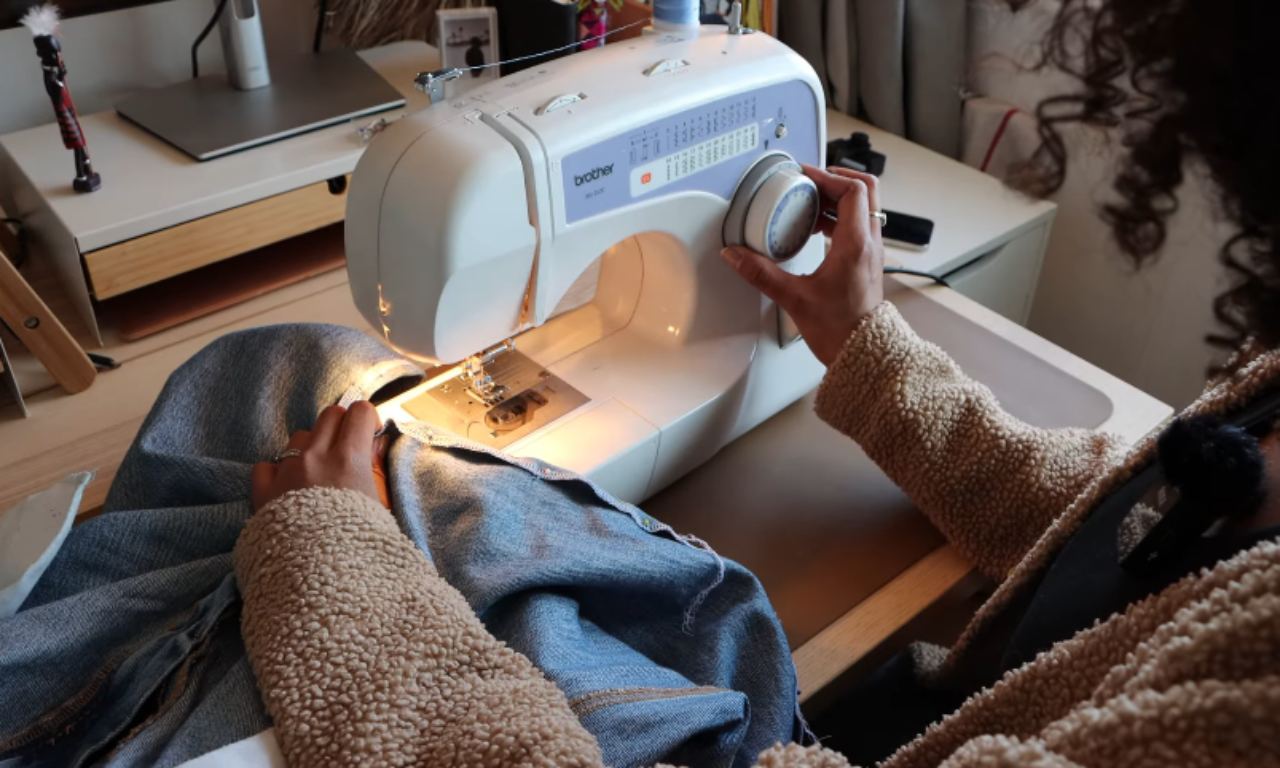 A person uses a white sewing machine to sew a new seam on denim fabric, a key step in the DIY project of turning jeans into a skirt.