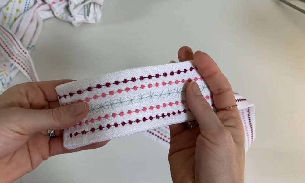 Two hands holding a finished white fabric belt, showcasing rows of decorative stitches in pink, burgundy, and light green made on a sewing machine.