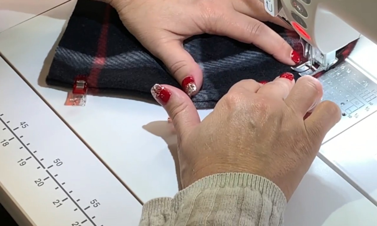A close-up of hands with red nails guiding black and red plaid fleece fabric under the presser foot of a sewing machine.