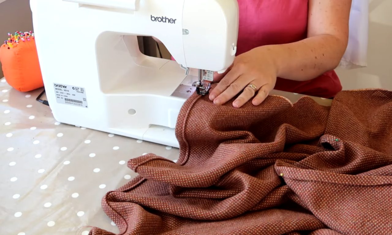 A person sewing a bench seat cushion cover on a white Brother sewing machine. Their hands are guiding the textured, rust-colored fabric under the needle, with an orange pincushion visible in the background.