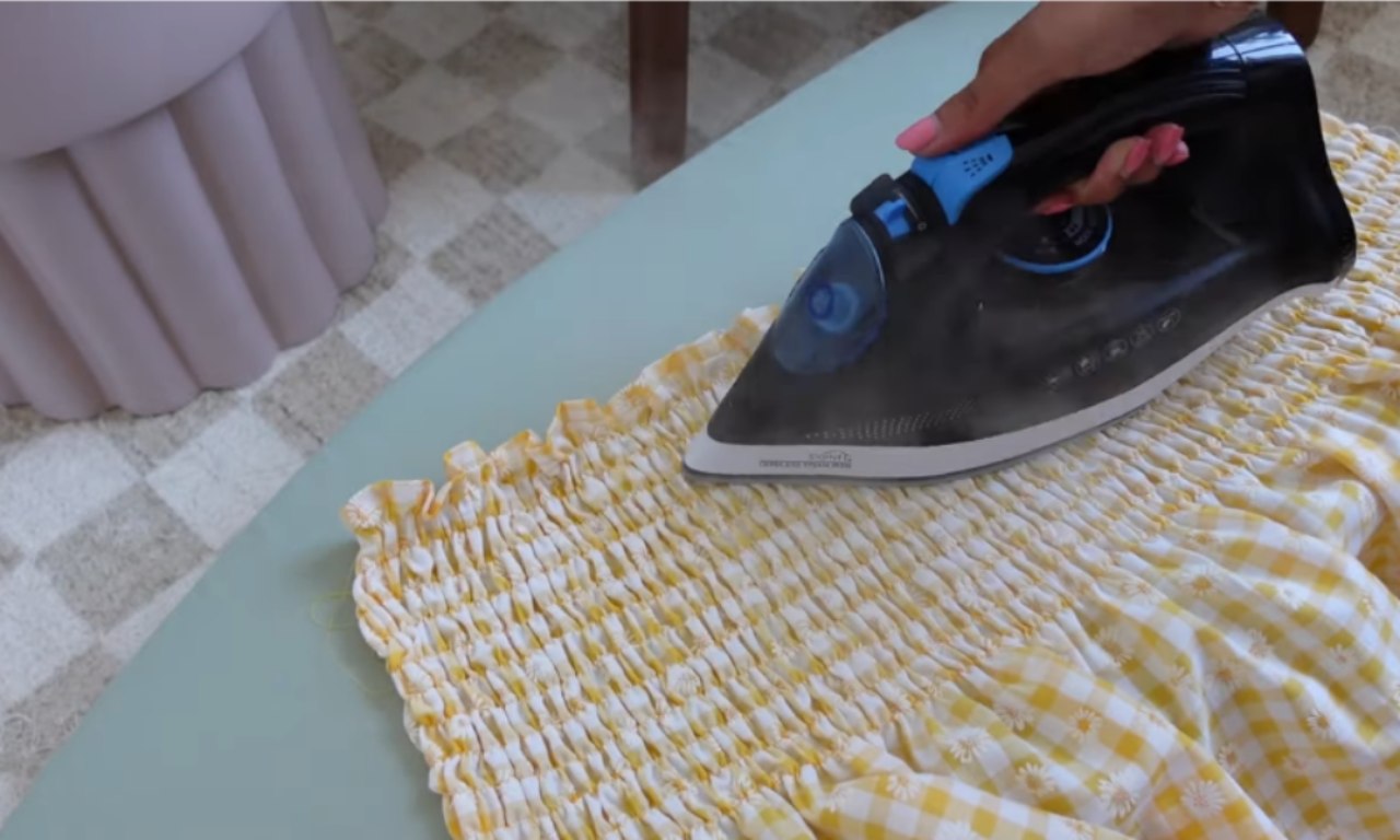 A hand using a steam iron to set the shirring on the bodice of a yellow gingham dress, making the elastic gathers shrink and tighten.