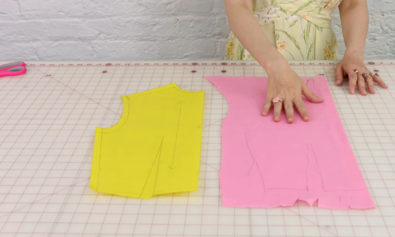 A person lays a pink fabric piece next to its yellow paper sewing pattern on a gridded cutting mat.