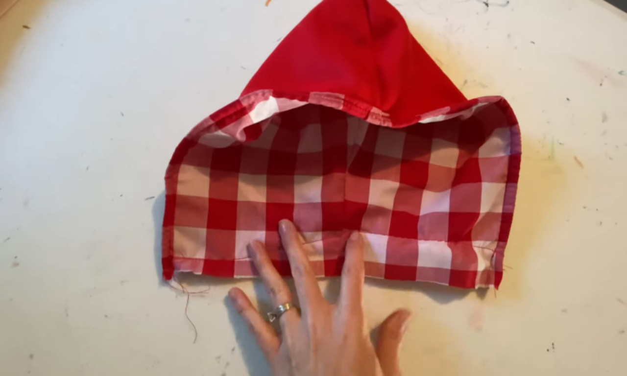 A completed hood for a kids cape, lying on a white surface. The outer fabric is solid red, and the inner lining is a red and white checkered pattern.